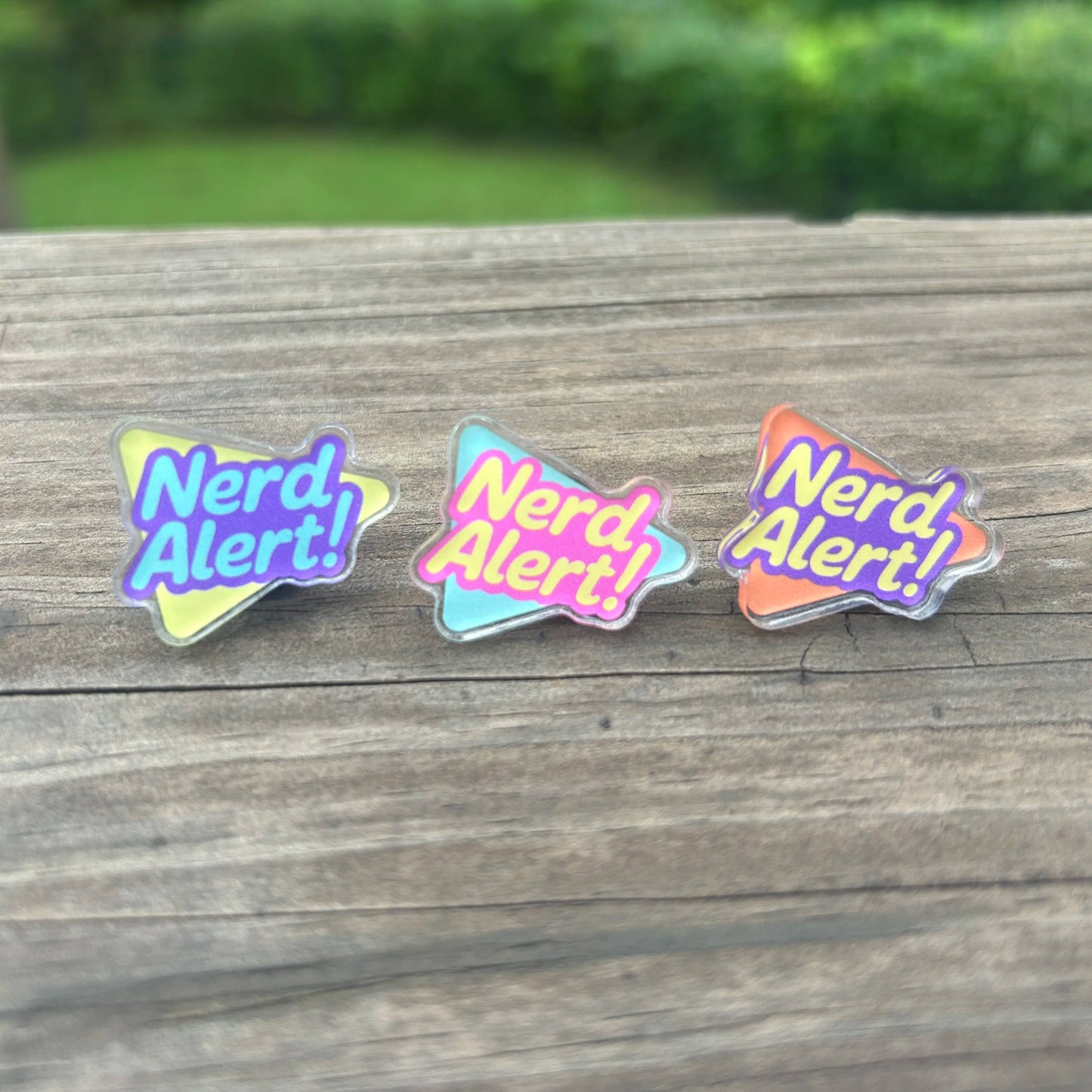 Three 'Nerd Alert!' pins on a wooden surface with a blurred green background