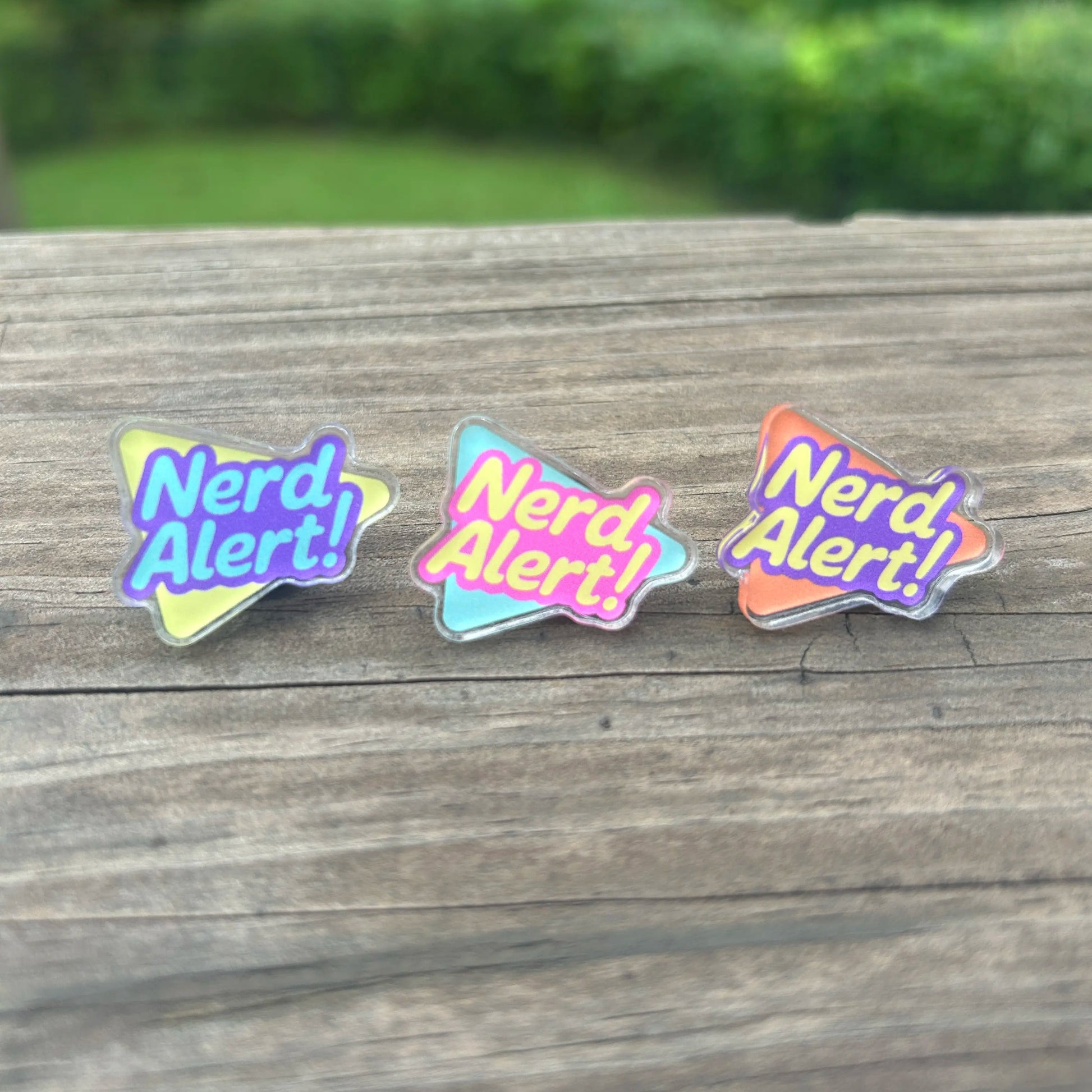 Three 'Nerd Alert!' pins on a wooden surface with a blurred green background